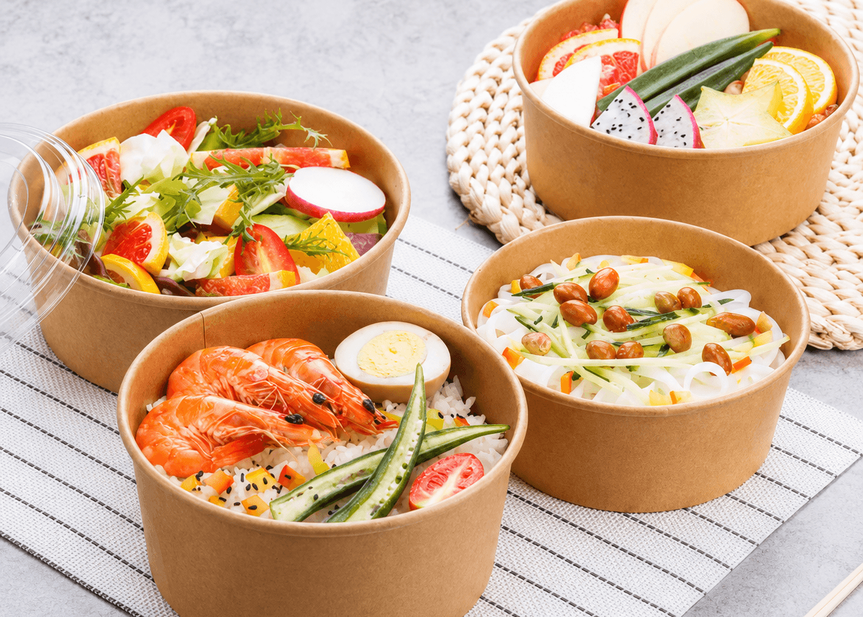Four round kraft paper take-out containers filled with different prepared foods, arranged on a striped mat over a gray surface. The containers are shown with colorful salads, shrimp and rice, and fruit slices.