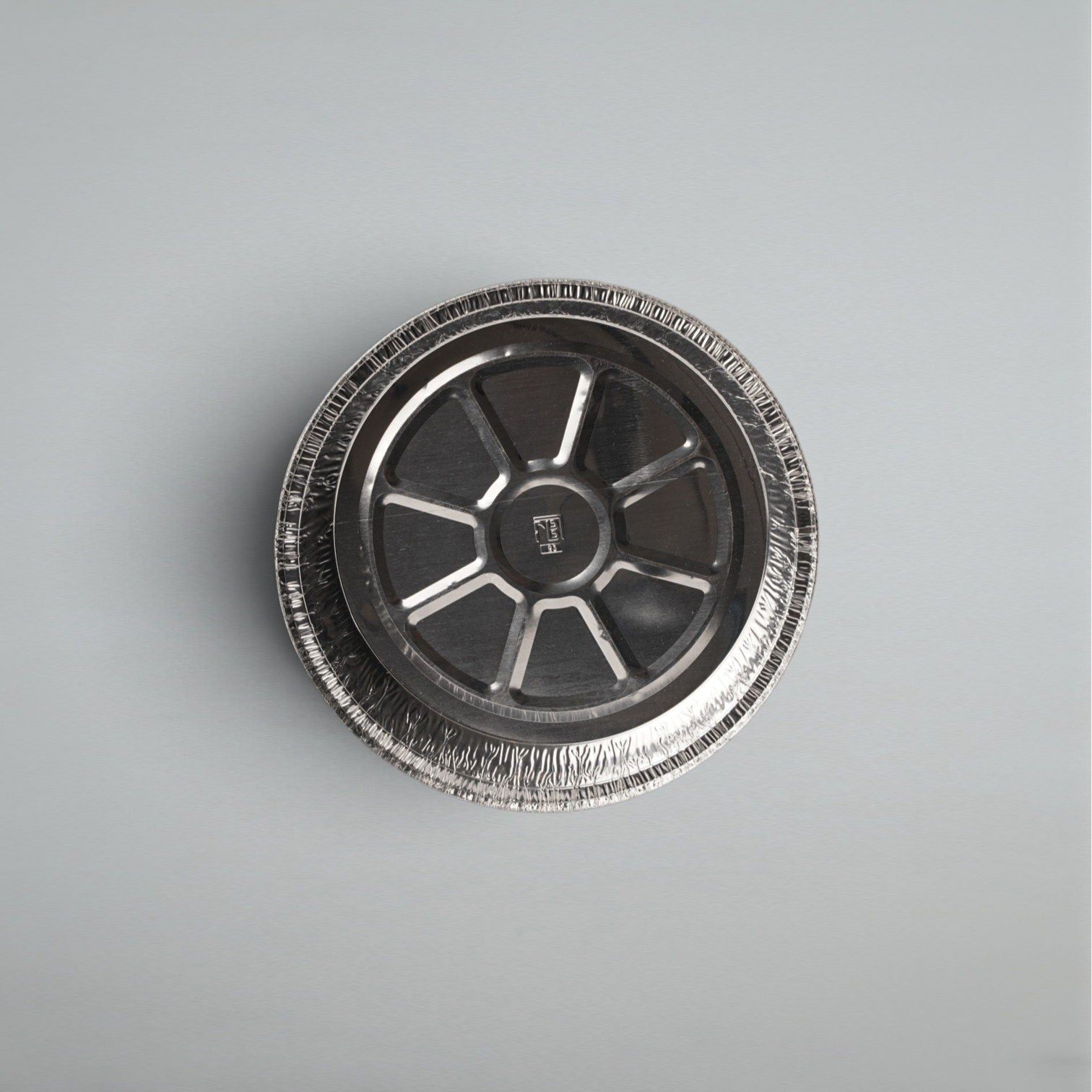 A round aluminum foil pie tin viewed from directly above, showing its textured crimped rim and metallic interior, set against a plain light gray background.