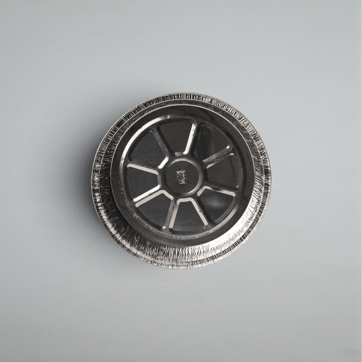 A round aluminum foil pie tin viewed from directly above, showing its textured crimped rim and metallic interior, set against a plain light gray background.