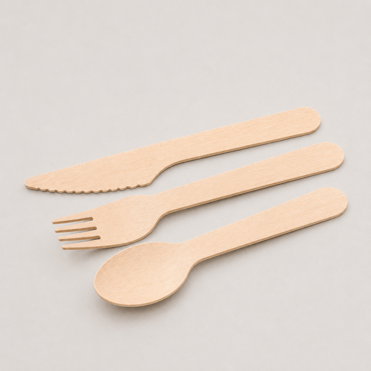 Three light natural wood disposable cutlery pieces, including a spoon, fork, and knife, arranged diagonally on a neutral light gray background.