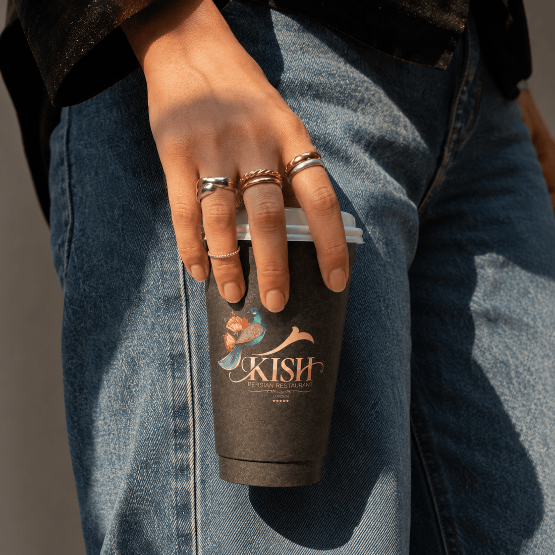 Kish Persian Restaurant branded black takeaway coffee cup with lid, held by a customer in a warm lifestyle setting.