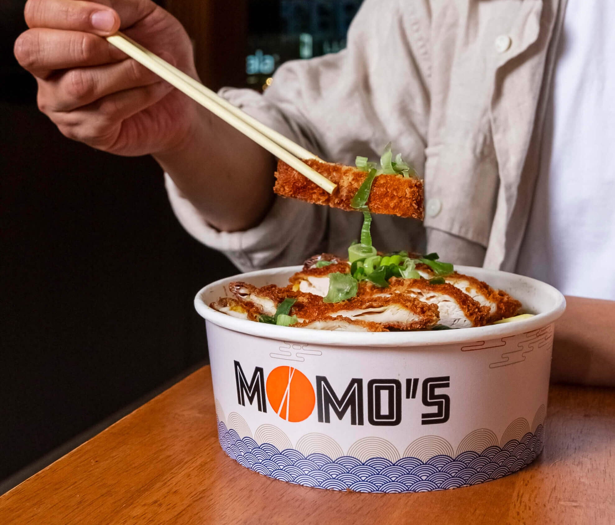 Momo’s branded takeaway food bowl filled with crispy chicken katsu and fresh garnish, with a customer eating using chopsticks.