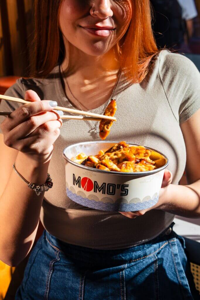 Momo’s branded takeaway food bowl filled with an Asian-inspired meal and held by a customer eating with chopsticks.